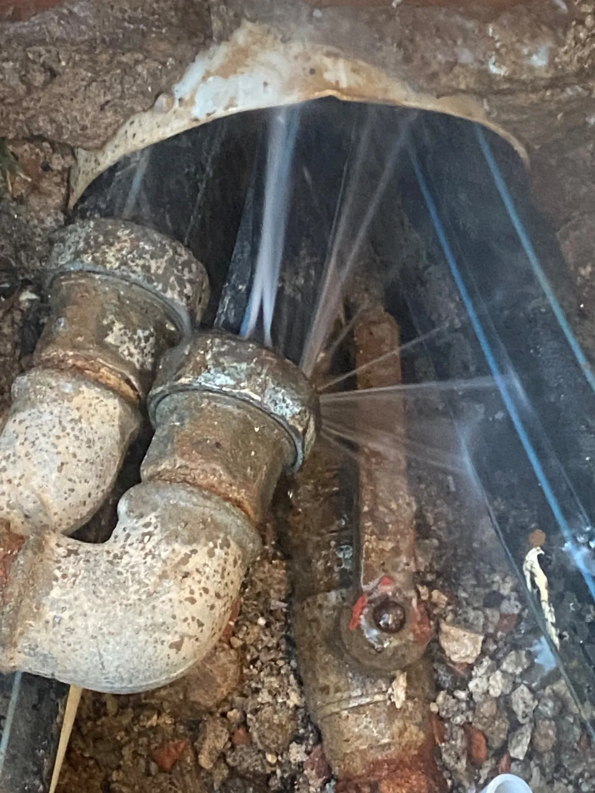 Getti d'acqua escono dal dado di un raccordo metallico arrugginito in uno scavo, accanto a tubi neri a righe blu