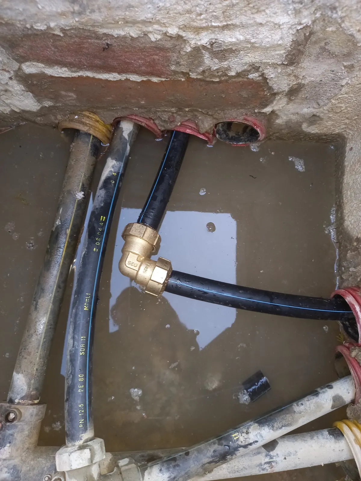 Pozzetto con acqua sul fondo: tubo in polietilene nero a riga blu collegato con un gomito in ottone vicino alle guaine corrugate rosse