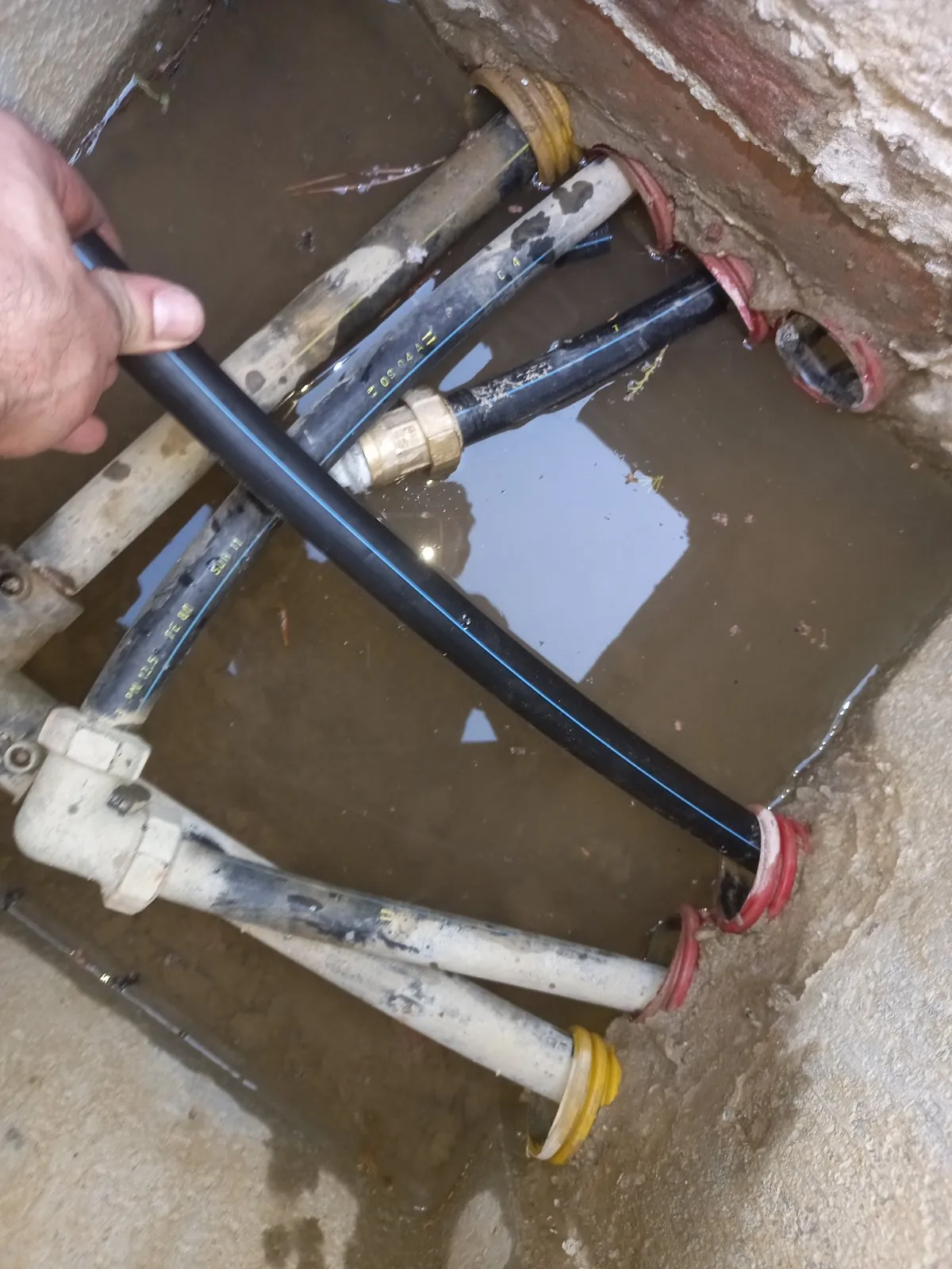 Mano che infila un tubo in polietilene nero a riga blu nella guaina corrugata rossa, in un pozzetto con acqua sul fondo