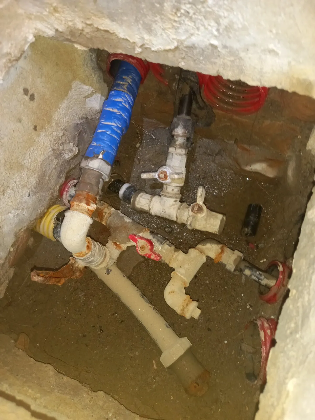 Pozzetto con valvole, raccordi arrugginiti, tubo rivestito blu e guaine corrugate rosse e gialle da cui escono le tubazioni