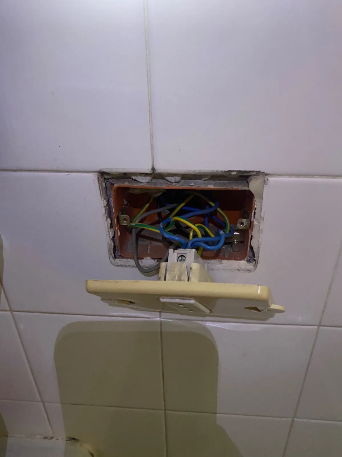 Vecchia presa con placca avorio staccata dalla scatola incassata tra le piastrelle, con fili blu, giallo-verdi e grigi a vista